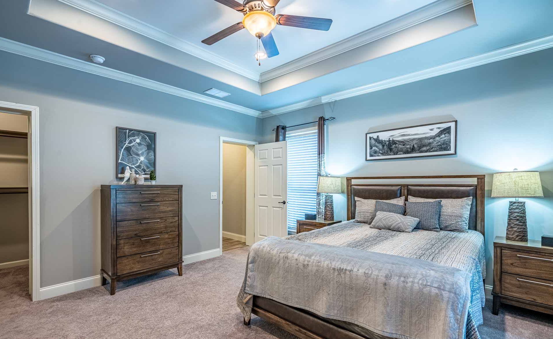 Spacious bedroom with a ceiling fan, soft blue walls, and a plush carpet. Features a large bed, wooden dresser, nightstands, and serene artwork, creating a calm, inviting atmosphere.