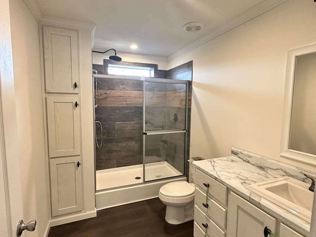 Modern bathroom with a glass-enclosed shower, dark wood accent wall, white cabinets, marble countertop, and bright lighting, conveying a clean and calming ambiance.