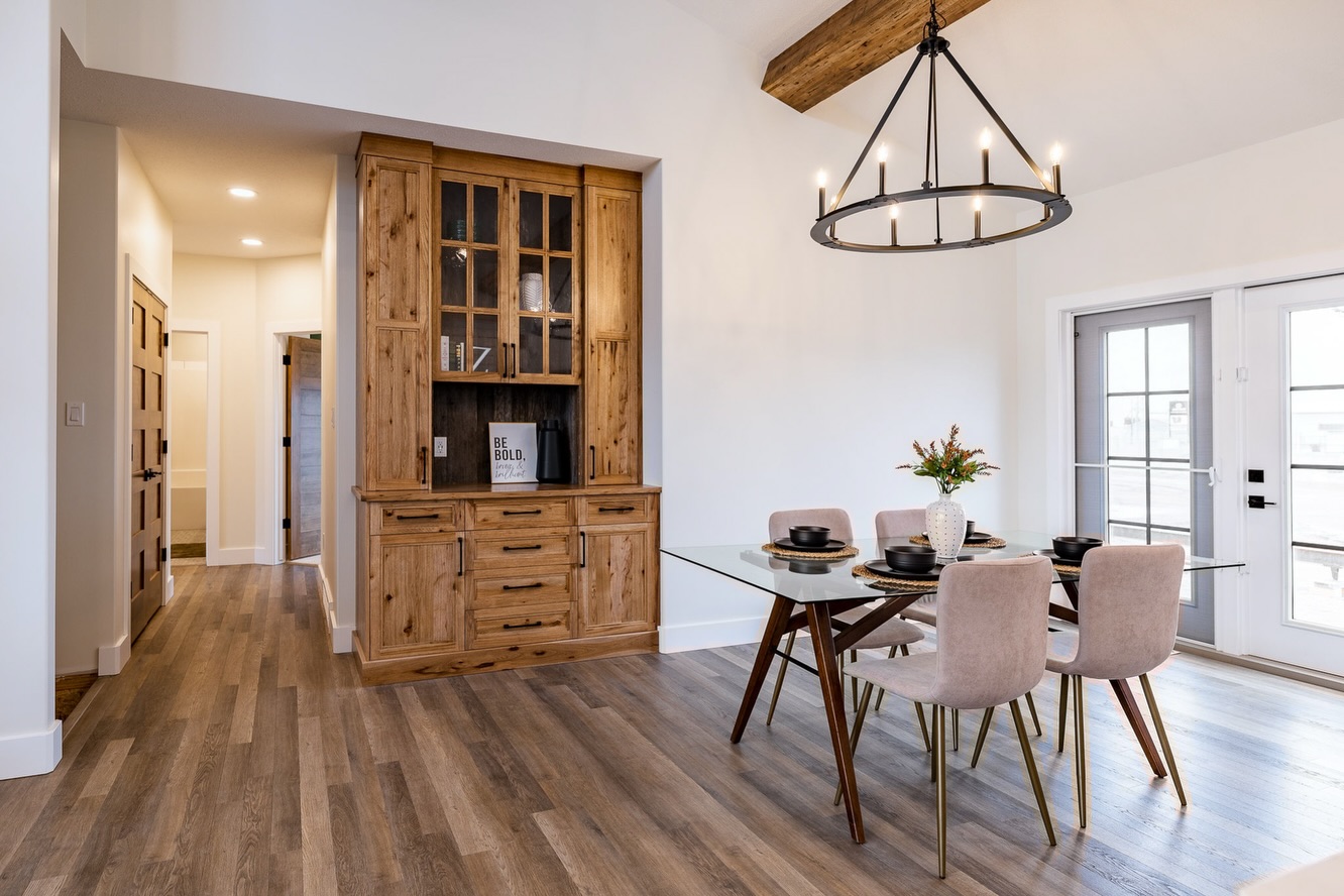 Modern dining room with wooden floors, a rustic wooden hutch, and a glass table surrounded by pink chairs. A large chandelier hangs above, and French doors let in natural light.