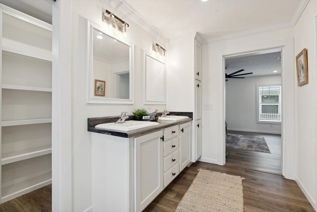 Spacious luxurious master bathroom and walk-in closet hallway in modern modular home featuring double white undermount sinks on dark wood-grain quartz countertop, white shaker-style vanity cabinets with black hardware, large rectangular mirrors with integrated sconce lighting, open built-in white shelving for storage, crown molding and textured ceiling, light gray luxury vinyl plank flooring with neutral jute area rug, arched doorway leading to bright bedroom with ceiling fan and large windows, elegant transitional farmhouse-style primary ensuite interior view in new manufactured home