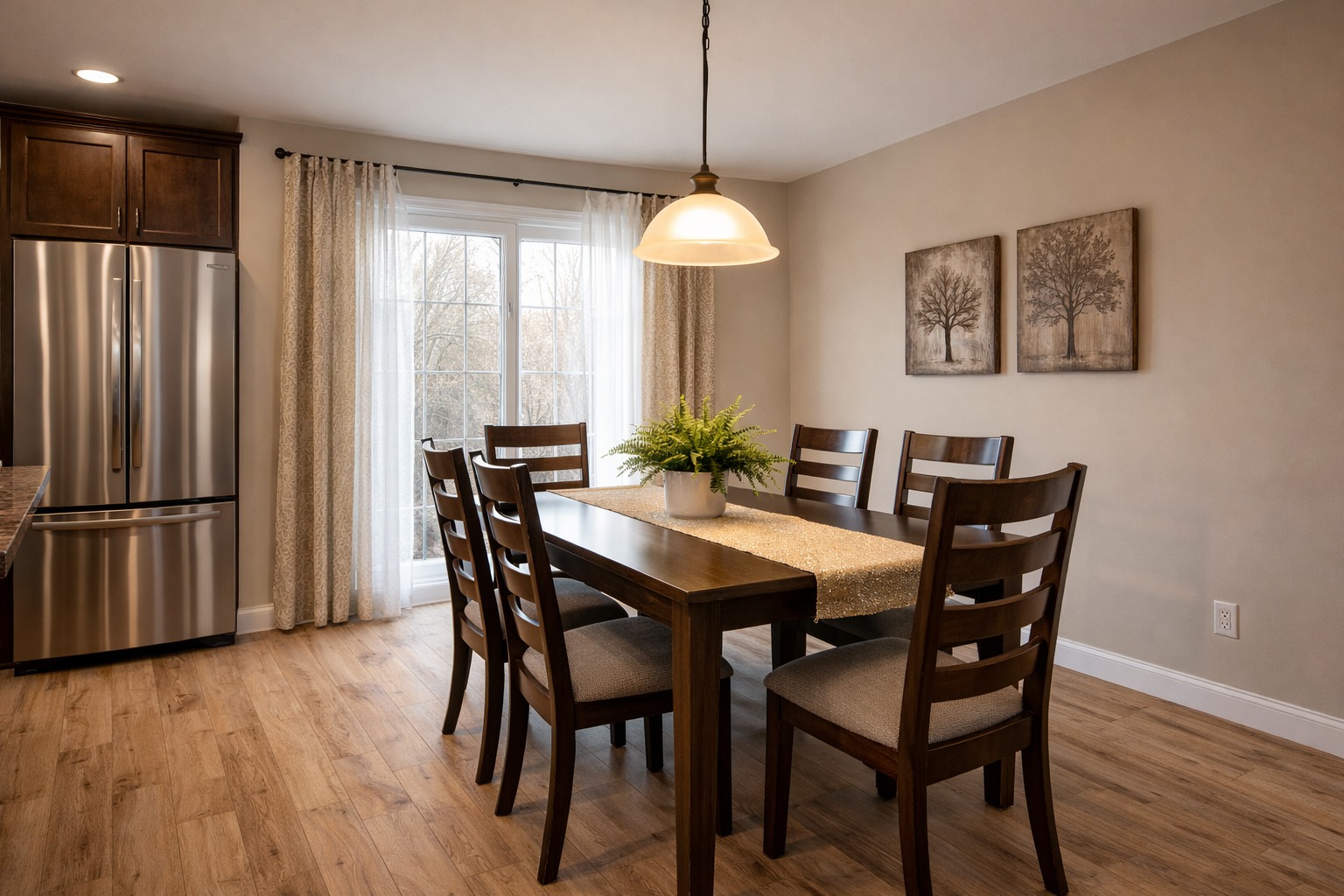 A cozy dining room with a wooden table set for six, a potted fern centerpiece, and a pendant light overhead. Natural light filters through sheer curtains on a window. Neutral-toned walls feature two tree artworks, and a stainless steel fridge is visible in the background, creating a warm, inviting atmosphere.