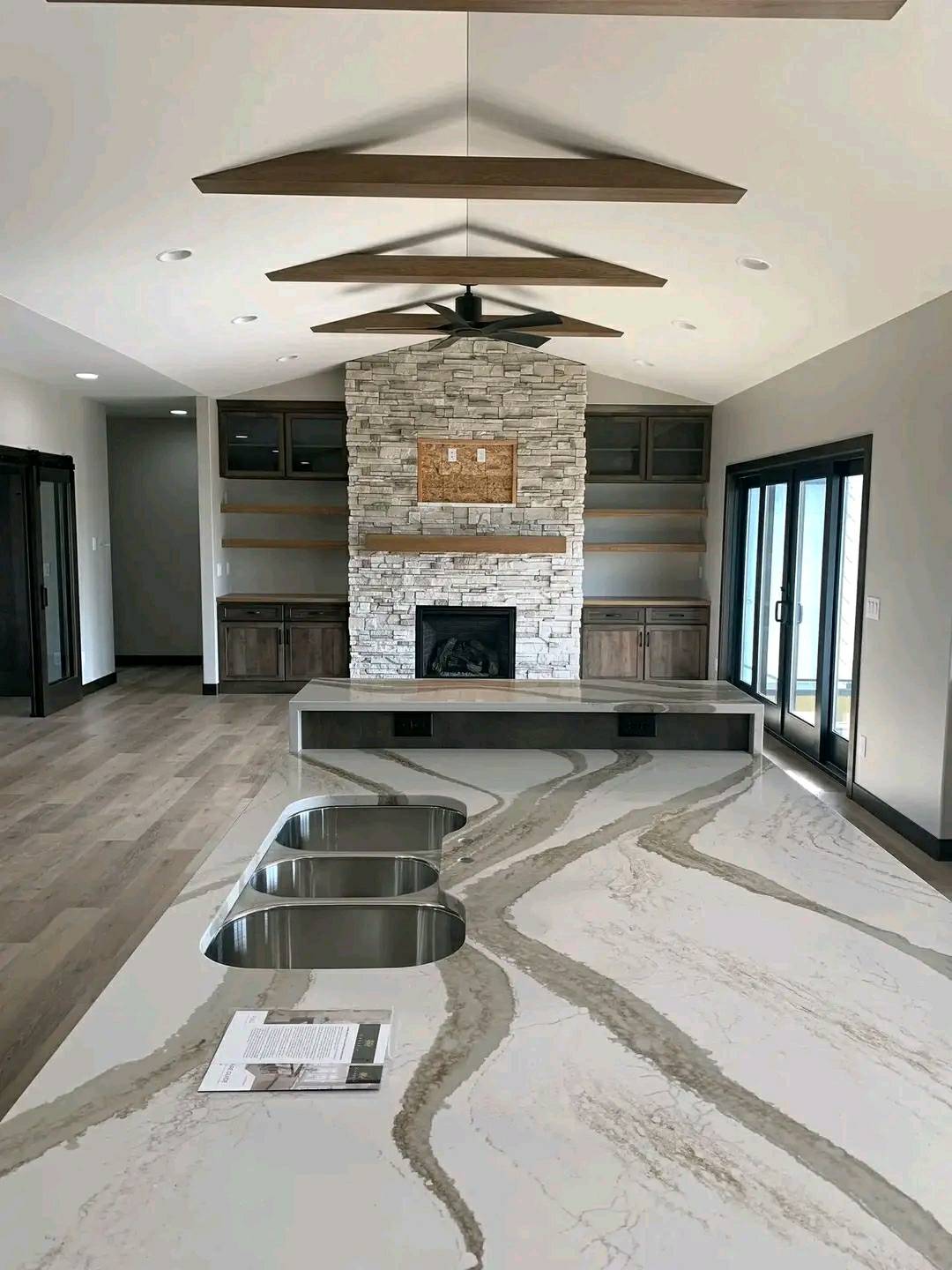 Modern kitchen interior featuring a large marble island with swirling patterns, dual sinks, wooden ceiling beams, stone fireplace, and sliding glass doors.