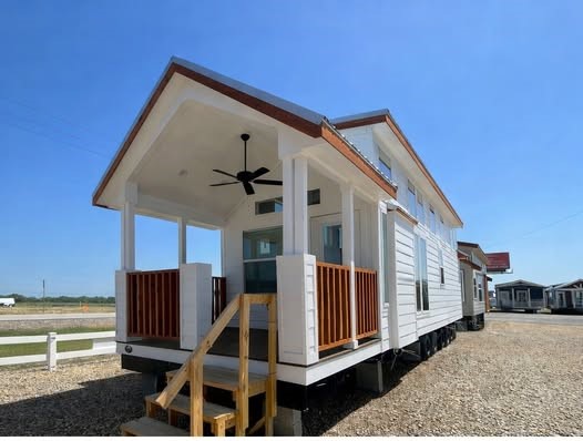 A modern tiny house on wheels with white siding and wood accents, featuring a small porch with a ceiling fan. It's set on a gravel lot under a clear blue sky.