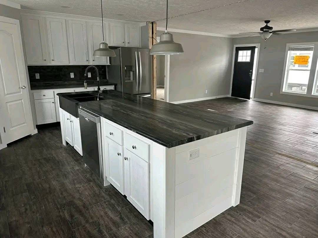 Modern kitchen with a dark wooden island, white cabinets, stainless steel appliances, and pendant lights. Open, bright space with wood floors.