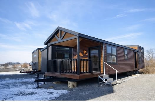 A wooden tiny house on wheels with a gabled roof and covered porch sits on a snowy landscape under a clear blue sky, conveying tranquility and mobility.