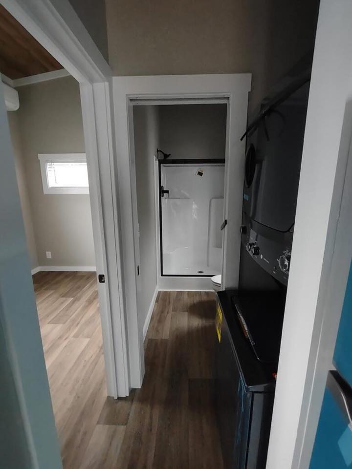 A narrow hallway with wood flooring leads to a small, white shower room. To the right, stacked laundry machines are visible, conveying functionality.
