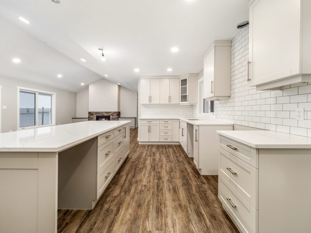 Luxurious open-concept gourmet kitchen in premium modern manufactured home featuring expansive white marble-look quartz countertops on large L-shaped island and perimeter cabinetry, white shaker cabinets with brushed nickel pulls, white subway tile backsplash, built-in stone accent electric fireplace with rustic brick surround, dark wood-look luxury vinyl plank flooring, vaulted tray ceiling with recessed LED lighting and modern pendant fixtures, floor-to-ceiling windows and sliding glass doors overlooking scenic water view, bright spacious airy great room kitchen layout in high-end 2025 double-wide modular home design