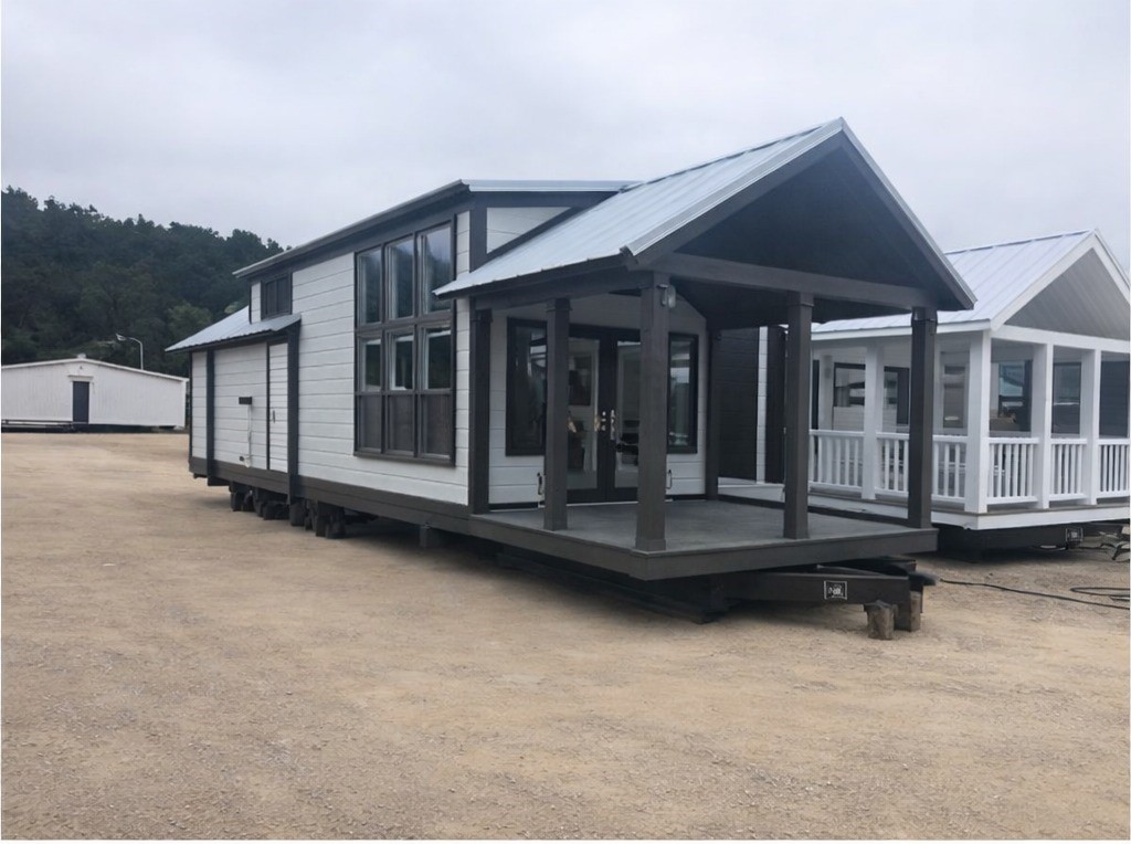 Modern gray and white tiny home on wheels with a covered porch, large windows, and a metal roof, parked outdoors on gravel with another tiny home visible to the right and a large white building in the background.