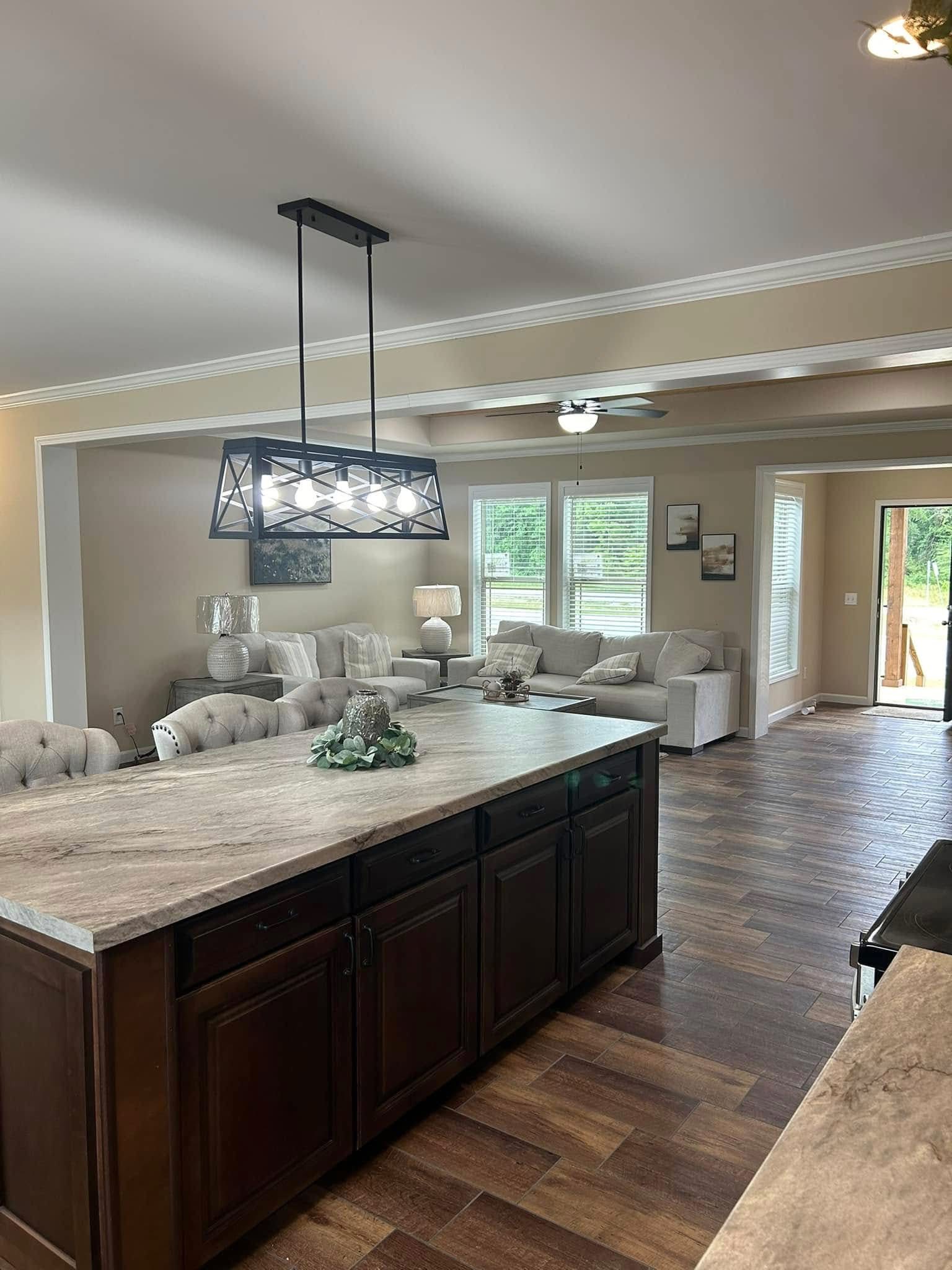 Spacious open-concept living area with a modern kitchen island featuring a granite countertop and dark wood cabinets, flowing into a living room with plush grey sofas, decorative lamps, and a large window overlooking a green outdoor space, all accented by rich wood-look tile flooring and a stylish geometric pendant light.