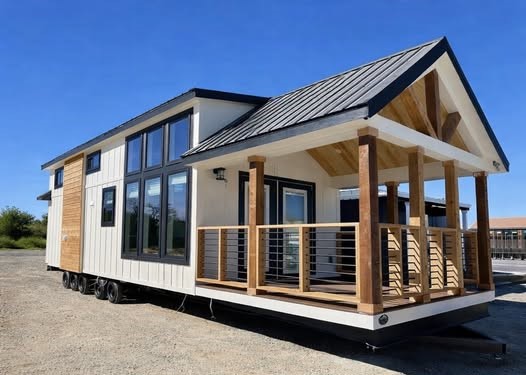 A modern tiny house on wheels with a gabled roof and wood accents. It features large windows and a cozy porch under a clear blue sky, conveying tranquility.