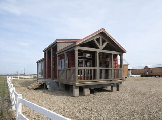 Compact rustic house on gravel lot, featuring a wooden porch and red siding. It’s surrounded by white fences and open sky, evoking simplicity and tranquility.