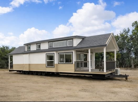 A modern tiny home on wheels sits on a dirt patch under a blue sky with clouds. The home features large windows, a small porch, and wood accents.