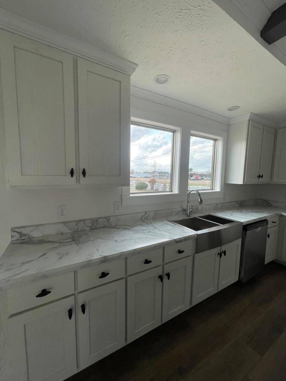 Bright kitchen with white cabinets, marble countertops, and a stainless steel sink. Large windows offer natural light, creating a fresh ambiance.