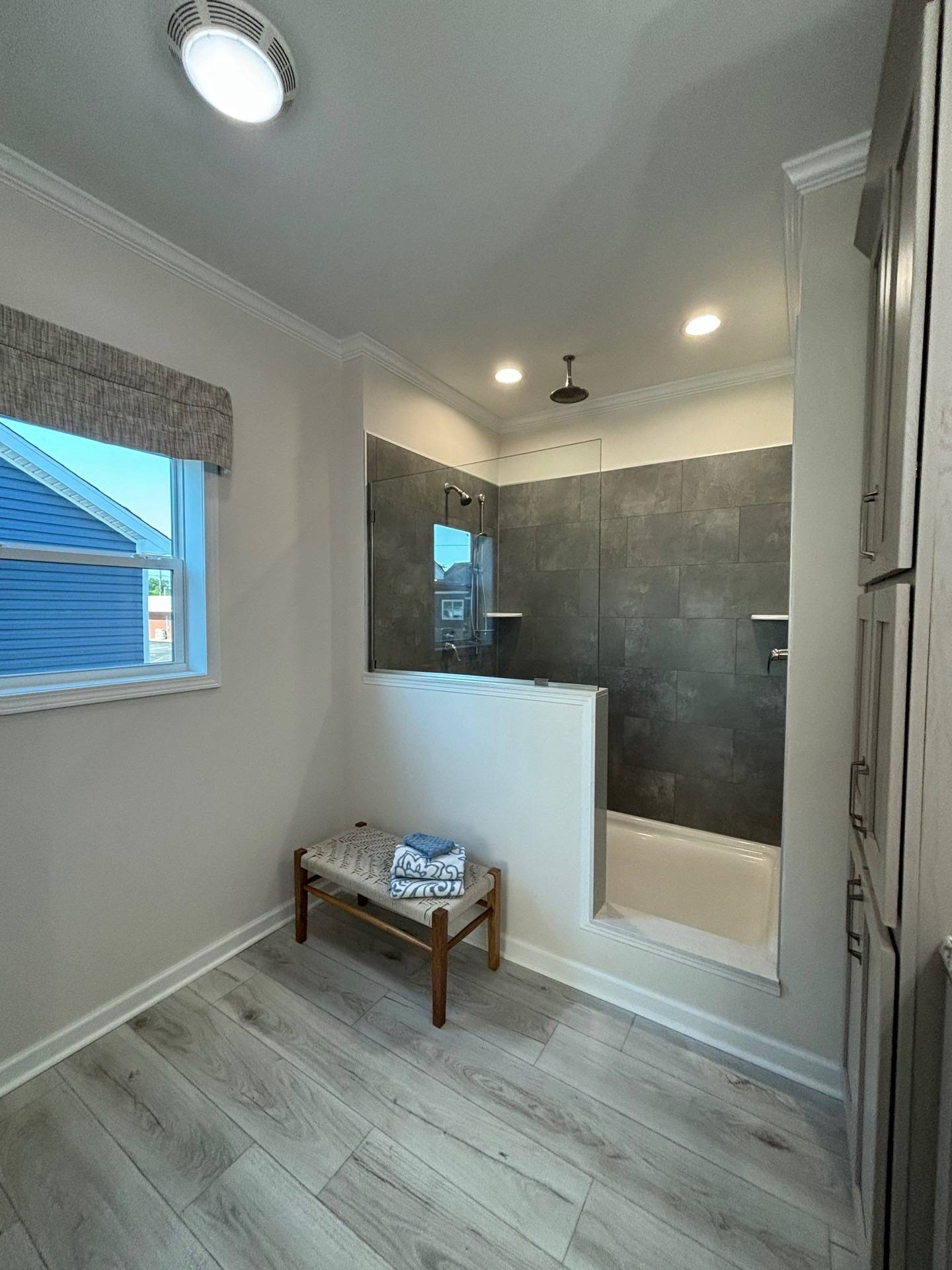 A modern bathroom with gray tile shower, glass door, and rain shower head. Light wood floor, window with gray shade, and a bench with towels. Calm and clean atmosphere.
