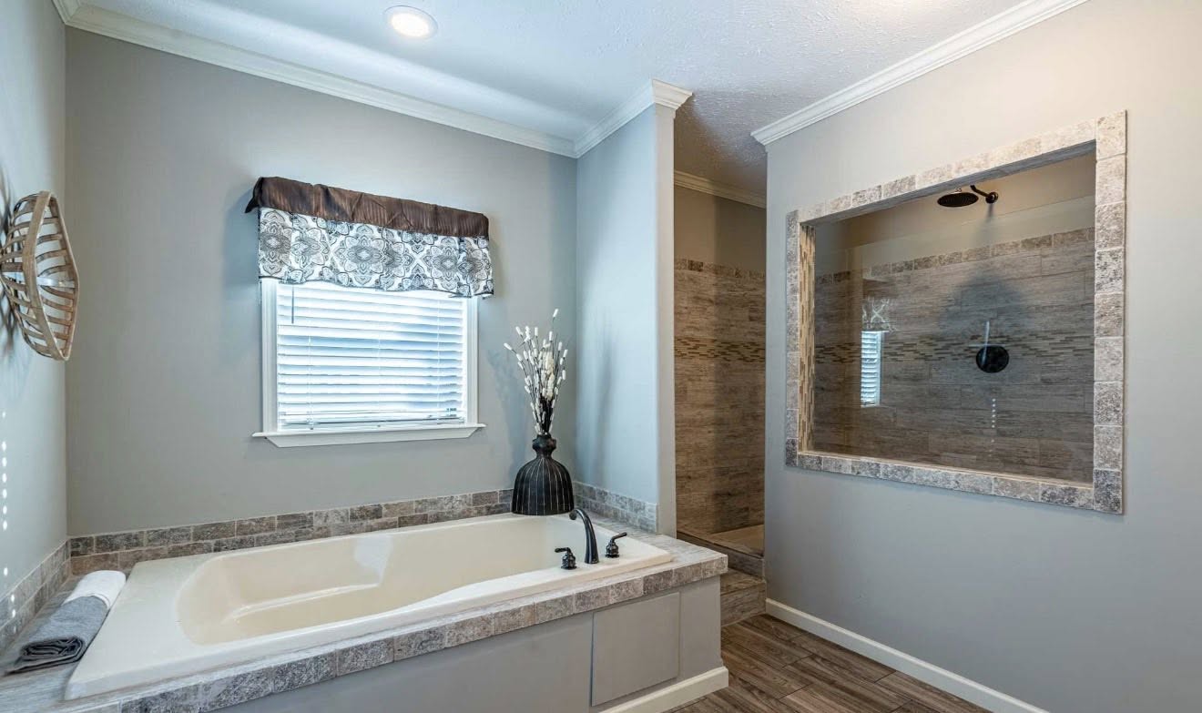 Modern bathroom with a large white tub, brown tiled trim, and a decorative vase beside it. A walk-in shower with brown tiles is in the background. Soft lighting creates a serene ambiance.