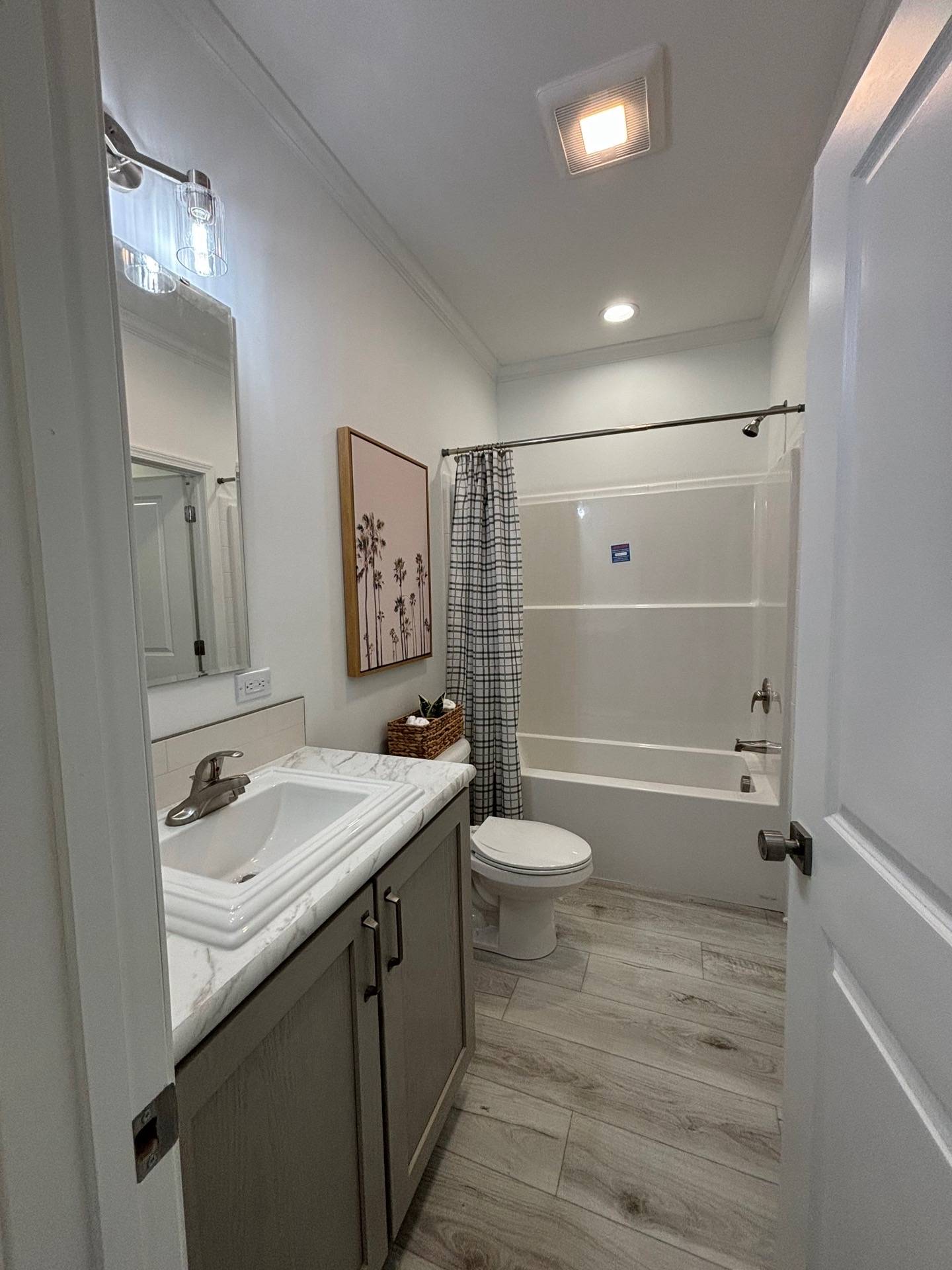 Narrow bathroom with wood-look flooring, featuring a white sink and cabinet on the left, a painting above, a toilet, and a bathtub with a plaid curtain.