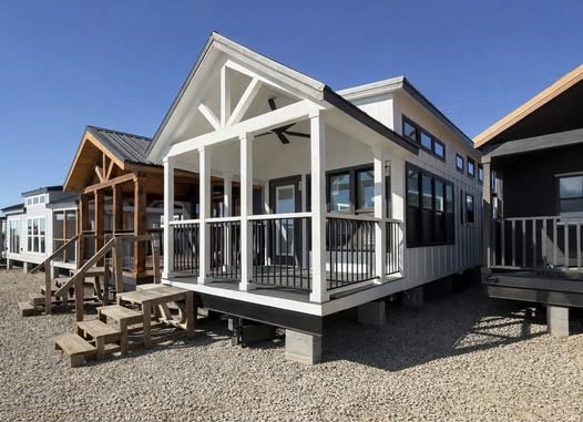 Row of small, modern cabins with gabled roofs and elevated decks, on a sunny day. The foreground cabin is white with dark trim and stairs.
