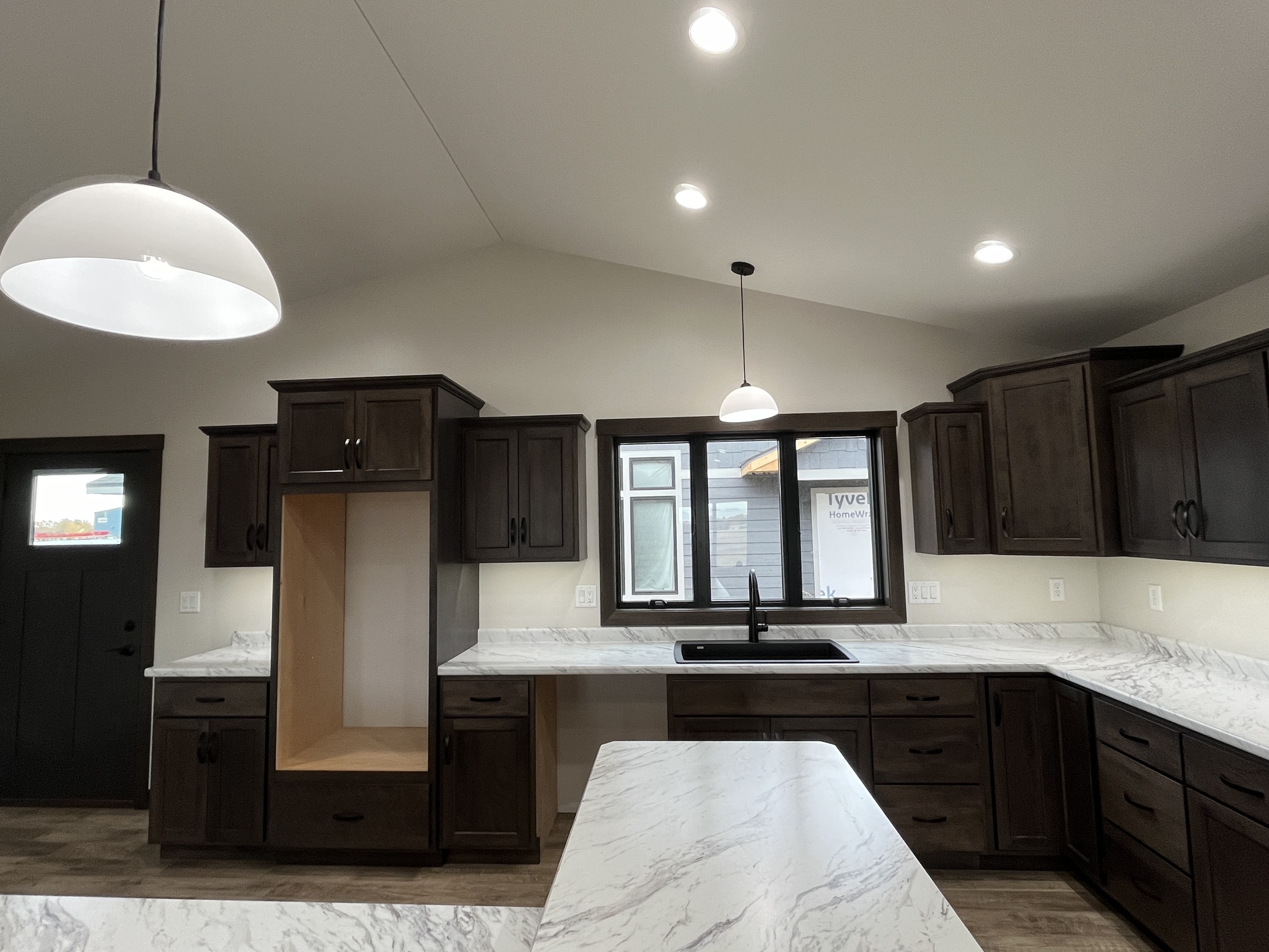 Modern luxury contemporary manufactured home kitchen featuring dark espresso-stained cabinetry, elegant white marble-patterned countertops, and a matching center island with a minimalist integrated sink. The sophisticated architectural design showcases a bright, vaulted ceiling with recessed LED lighting and stylish white dome pendant lights, while large black-trimmed energy-efficient windows provide a clear view of the professional construction environment. This "full rank" SEO-optimized space highlights premium wide-plank wood laminate flooring and a functional U-shaped layout, making it a perfect showcase for high-end prefab living, custom modular home floor plans, or modern permanent housing solutions designed with a sleek, upscale, and contemporary aesthetic.