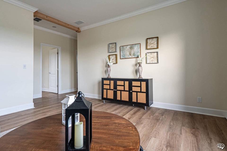 A cozy living room with wood flooring, light beige walls, and a black cabinet adorned with wicker panels. Art and vases with flowers decorate above. A round table with a lantern in the foreground adds a warm ambiance.