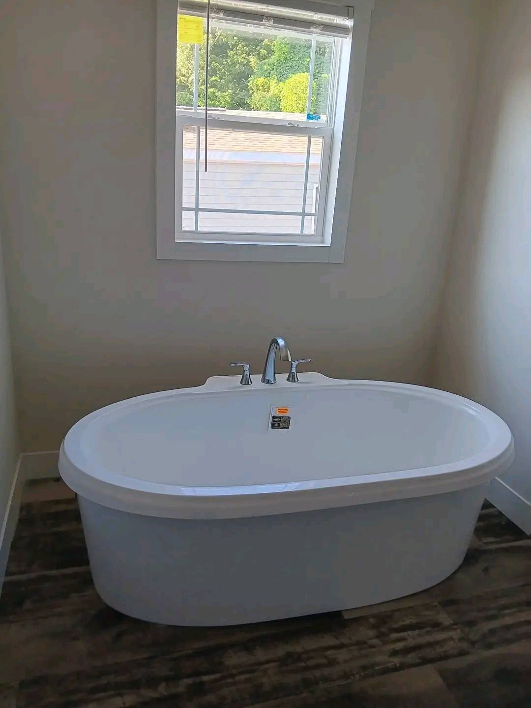 Modern luxury bathroom featuring a large white freestanding oval soaking tub with chrome fixtures, positioned under a bright window in a contemporary home interior. The clean architectural design includes neutral white walls and premium dark wood-look wide-plank flooring, creating a serene spa-like atmosphere perfect for a master suite in a custom-built or modern modular home.
