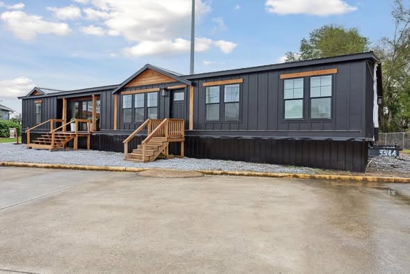 Modern stylish manufactured modular home exterior featuring sleek black vertical siding with natural wood trim accents, dual gable roofs, large multi-pane windows with wooden surrounds, welcoming covered front porch with dual wooden staircases and railings, stone and gravel foundation skirt, spacious single-story ranch-style bungalow facade under partly cloudy blue sky in residential setting
