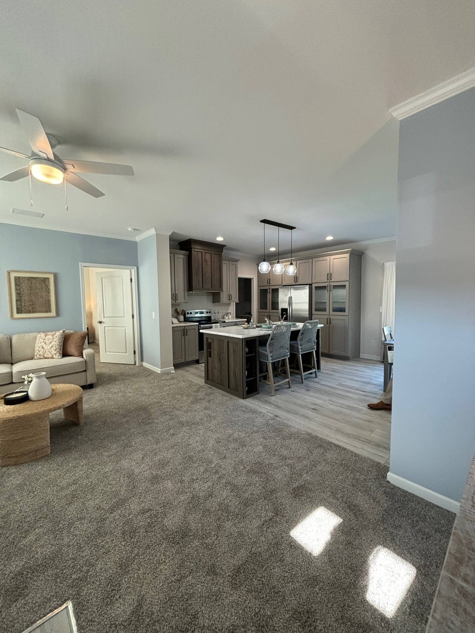 Spacious open-plan living room and kitchen with soft gray tones. Includes a cozy sofa, modern cabinets, island with bar stools, and decorative lighting.