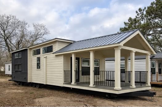 A modern tiny house with light siding and large windows. It features a covered porch with a dark metal roof and railings. The setting is a wooded area.