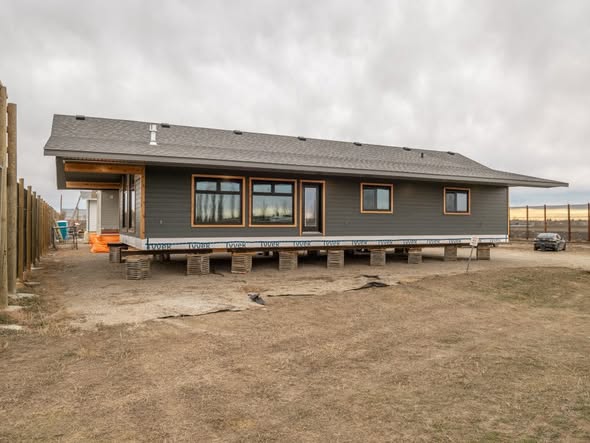 Elevated gray house on temporary supports, set on dry, grassy terrain under an overcast sky. Nearby, a fence and car are visible.