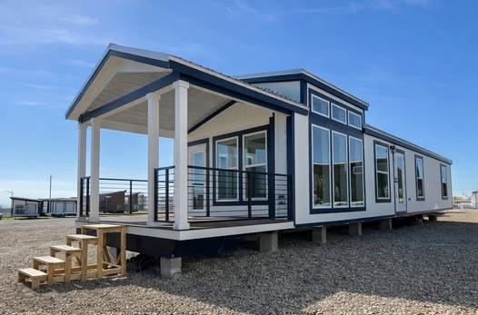Modern modular home with large windows and a small front porch on a gravel lot under a clear blue sky. Clean, minimalistic design conveys openness.