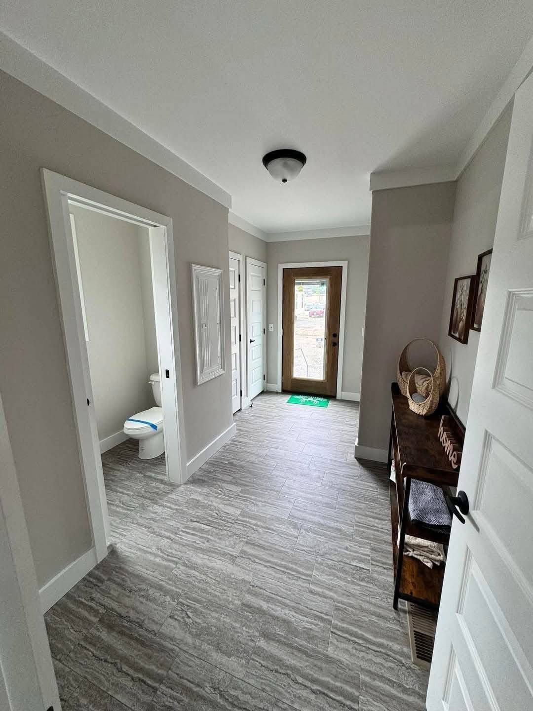 Bright hallway with light gray walls and tiled floor. On the left, a door opens to a small bathroom. The entrance at the end has a glass door. A wooden console on the right displays decorative items, creating a welcoming ambiance.