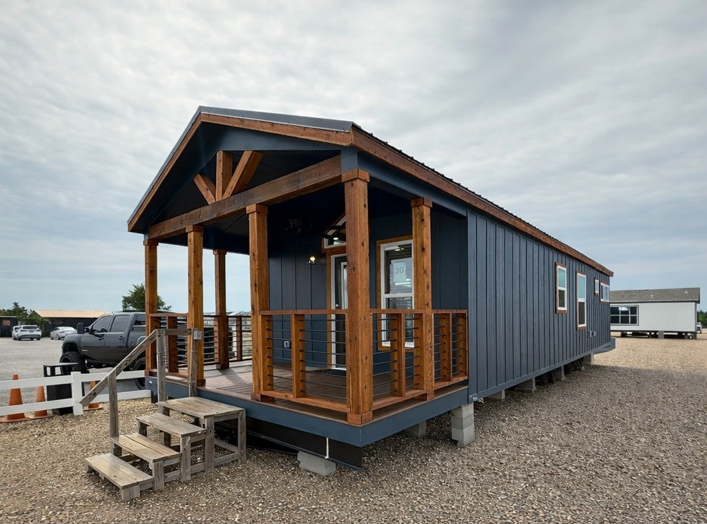 A small, dark gray modular home with wooden accents and a porch sits on a gravel lot under a cloudy sky. A truck is parked nearby.