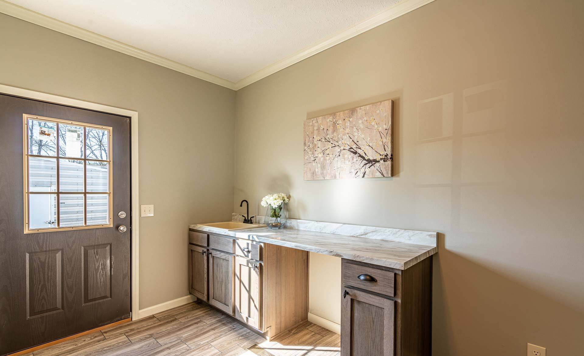 A small, bright room with beige walls features a dark wooden door with glass panes. A marble-topped counter with a sink and flowers sits to the right. A floral painting hangs above, adding a calm, inviting atmosphere.