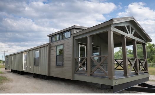 A modern mobile home with a wide front porch and gable roof, set on a dirt path. Gray siding, large windows, under a partly cloudy sky, creating a cozy look.