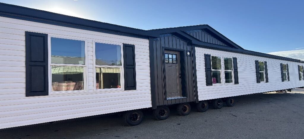 A white mobile home with black shutters and a dark wooden door sits on wheels under a clear blue sky, conveying a sense of mobility and potential.