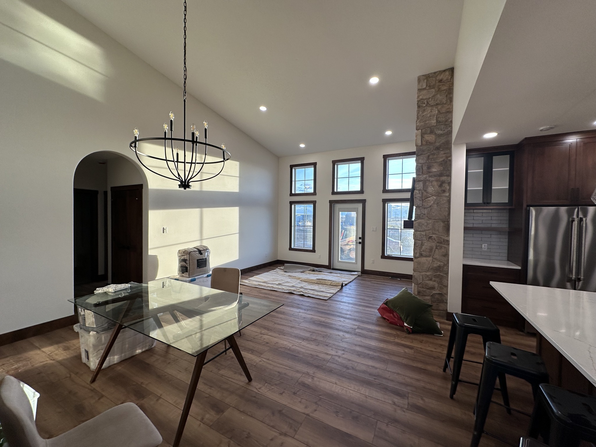 Spacious living area with high ceilings, hardwood floors, and large windows. A glass dining table sits under a modern chandelier. Stone accent wall and cozy tones.