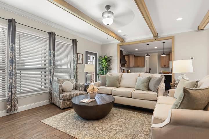 Bright living room with beige sofas, a patterned armchair, round wooden coffee table, and decorative rug. Wooden beams and pendant lights add warmth.