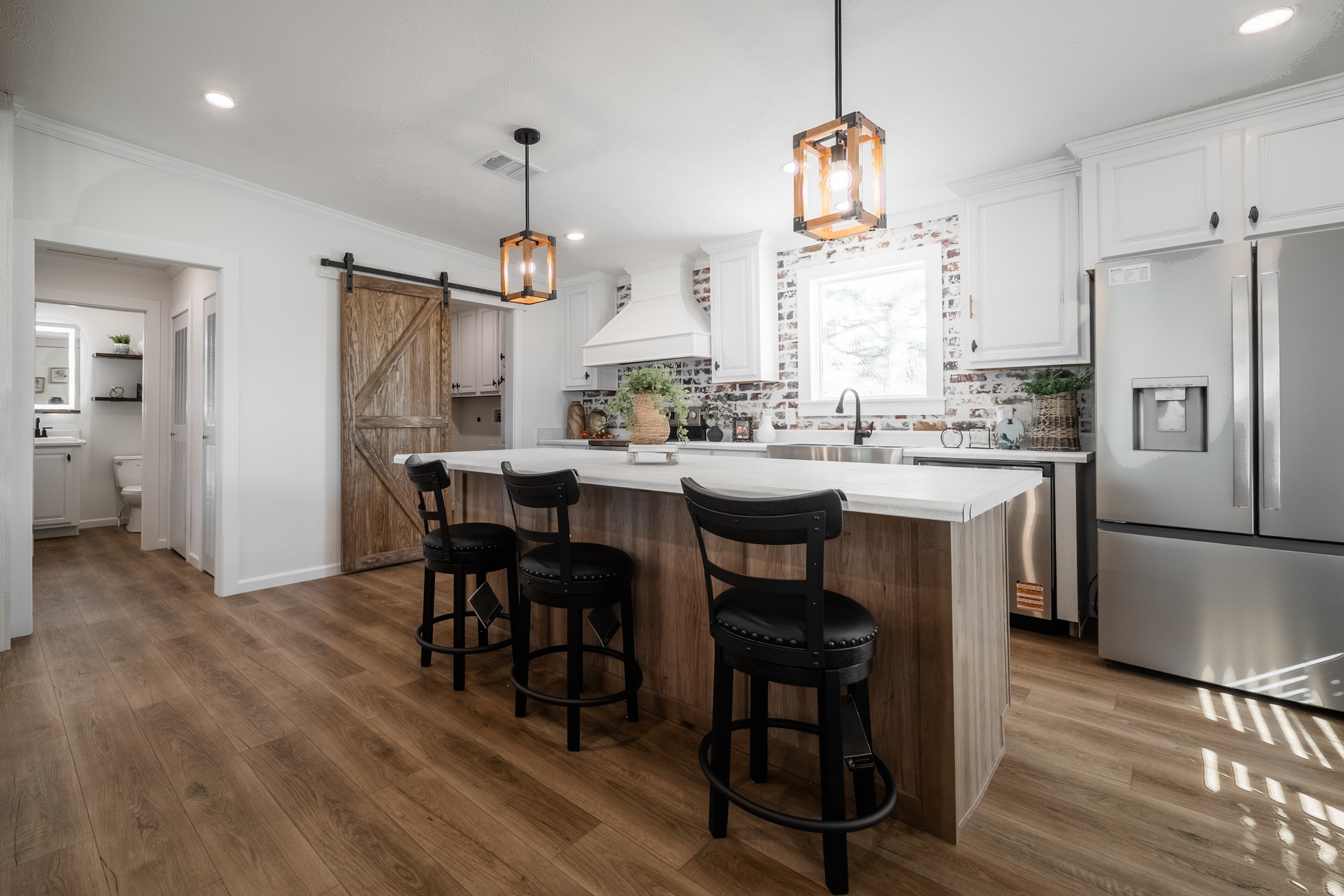 Modern kitchen with white cabinetry, a central island with three black chairs, and pendant lights. Barn door leads to a bathroom, creating a rustic touch.