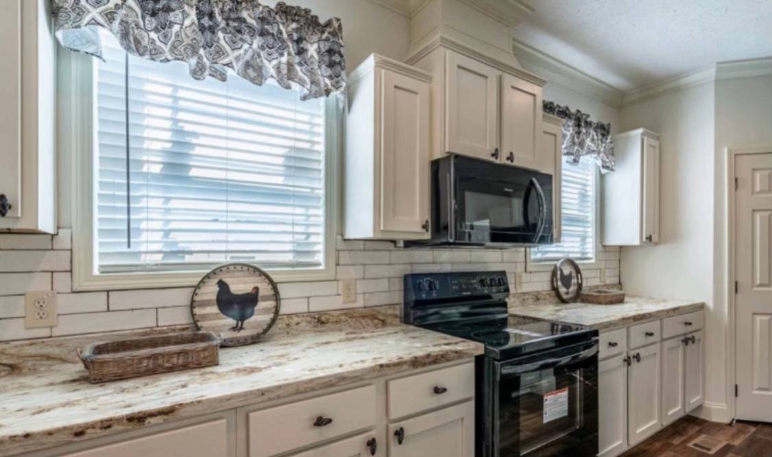 Bright kitchen with white cabinets, black appliances, and marbled countertops. Two windows with floral curtains let in natural light. Rooster decor adds charm.