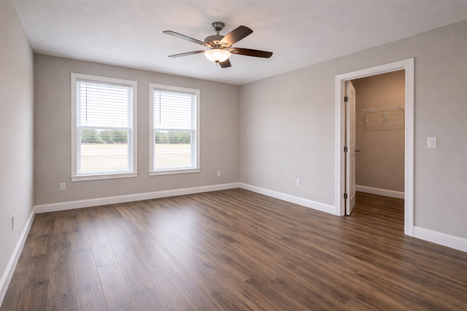 Empty room with light gray walls and wood flooring. Two large windows with blinds let in natural light. A ceiling fan is centered above. A door opens to a closet.