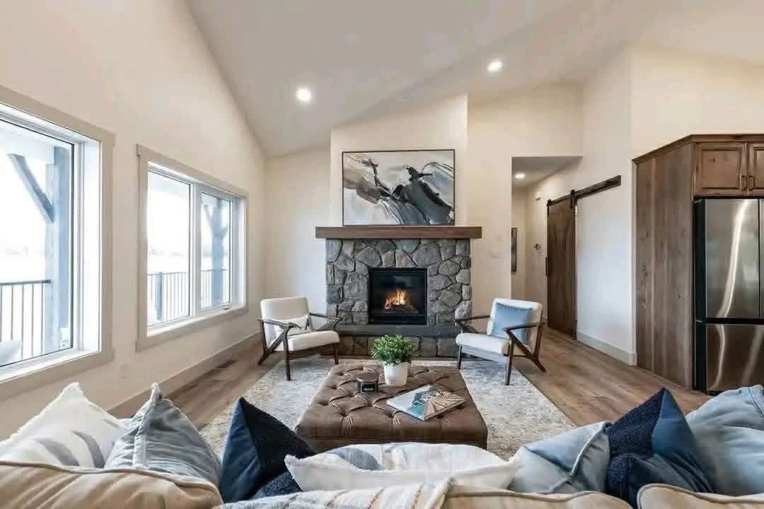 Cozy living room with a stone fireplace, modern abstract artwork above, two armchairs, a cushioned ottoman, and large windows. Warm, inviting ambiance.