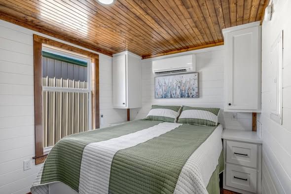 A cozy bedroom with a wooden ceiling and white paneled walls. The bed has a green and white quilt, flanked by built-in cabinets. A window to the left adds natural light.