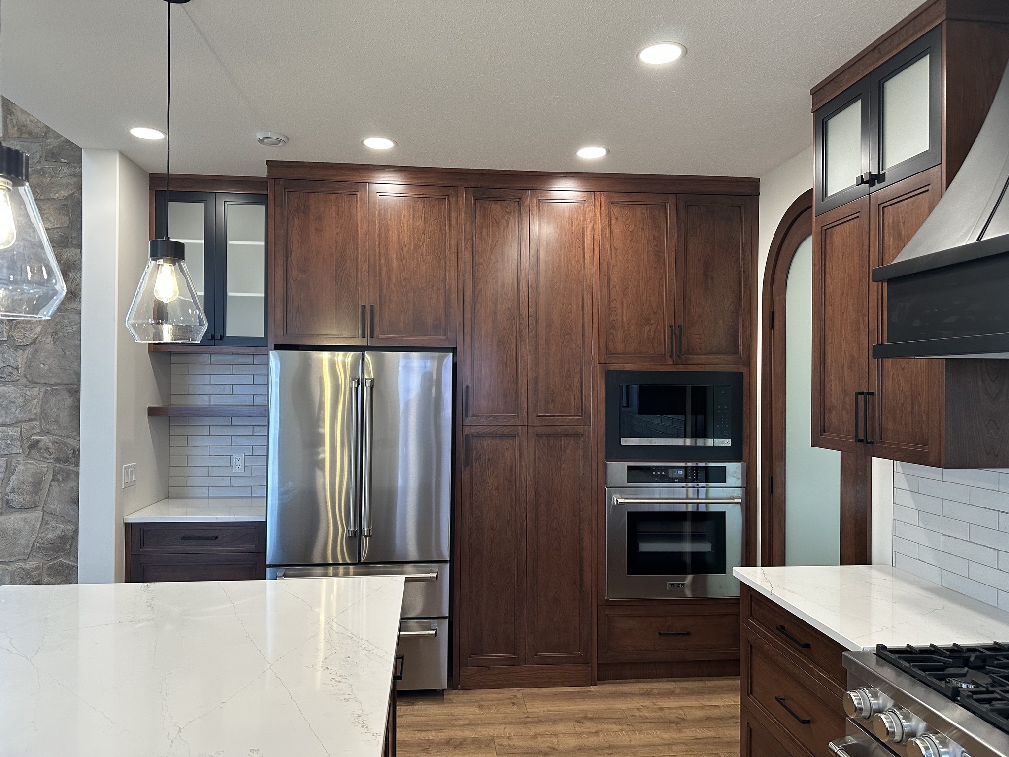 Modern kitchen with wooden cabinets, stainless steel fridge, and built-in ovens. Features pendant lights, white countertops, and a tile backsplash.