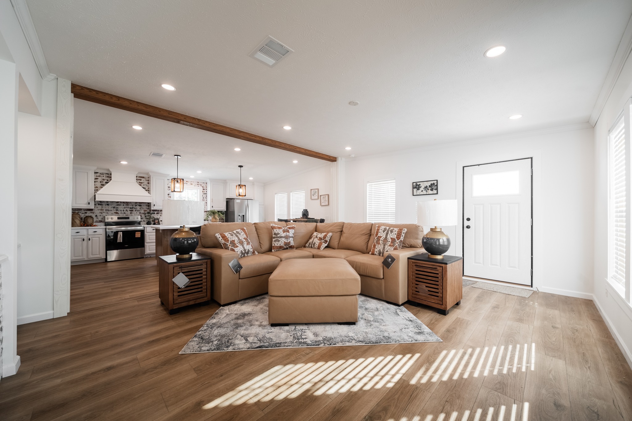 Open-concept living room with a beige sectional sofa on a gray rug, leading to a modern kitchen. Sunlight streams through large windows, creating a cozy atmosphere.
