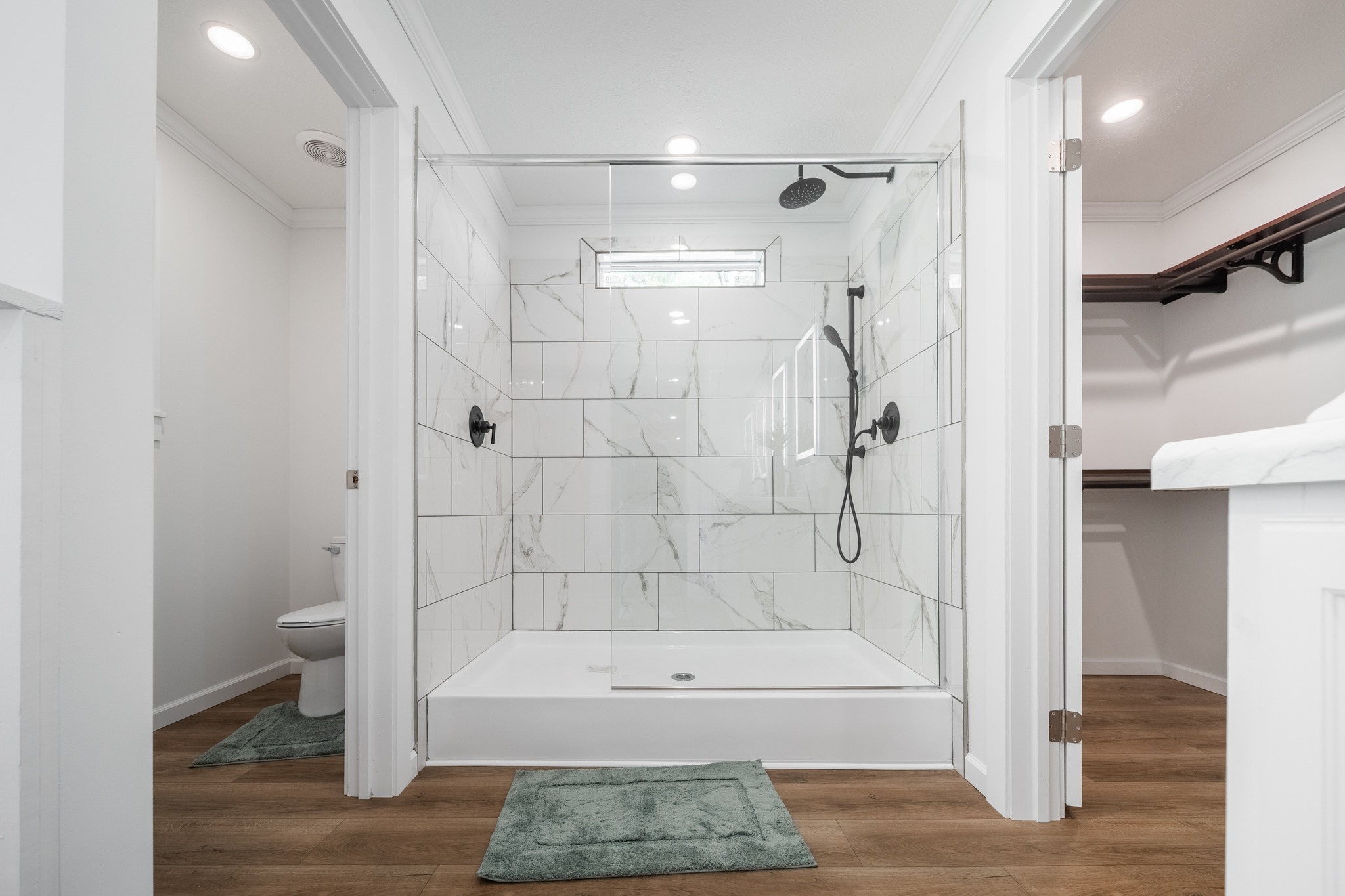 Modern bathroom with a large, glass-enclosed shower featuring marble tiles and a rainfall showerhead. Wooden floor, white walls, and soft green mats create a calm atmosphere.