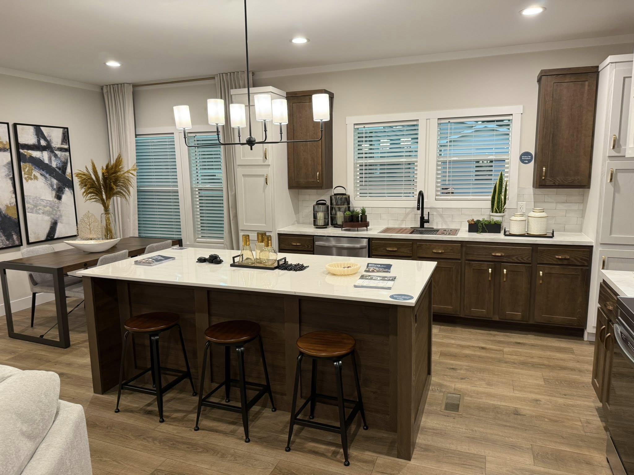 Modern kitchen with a central island featuring dark wood stools, elegant pendant lighting, wooden cabinets, and a mix of white and warm tones. Cozy ambiance.