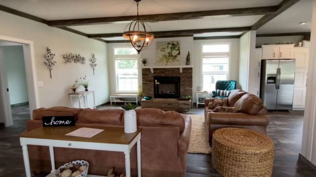 Cozy living room with brown leather sofas, stone fireplace, and rustic decor. Light pours through large windows, creating a warm, inviting atmosphere.