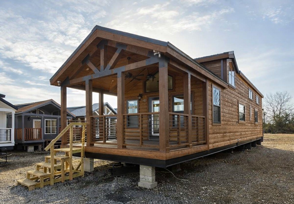A small wooden cabin with a gabled roof, elevated on pillars, features a front porch with railings and steps. The sky is partly cloudy, creating a serene atmosphere.