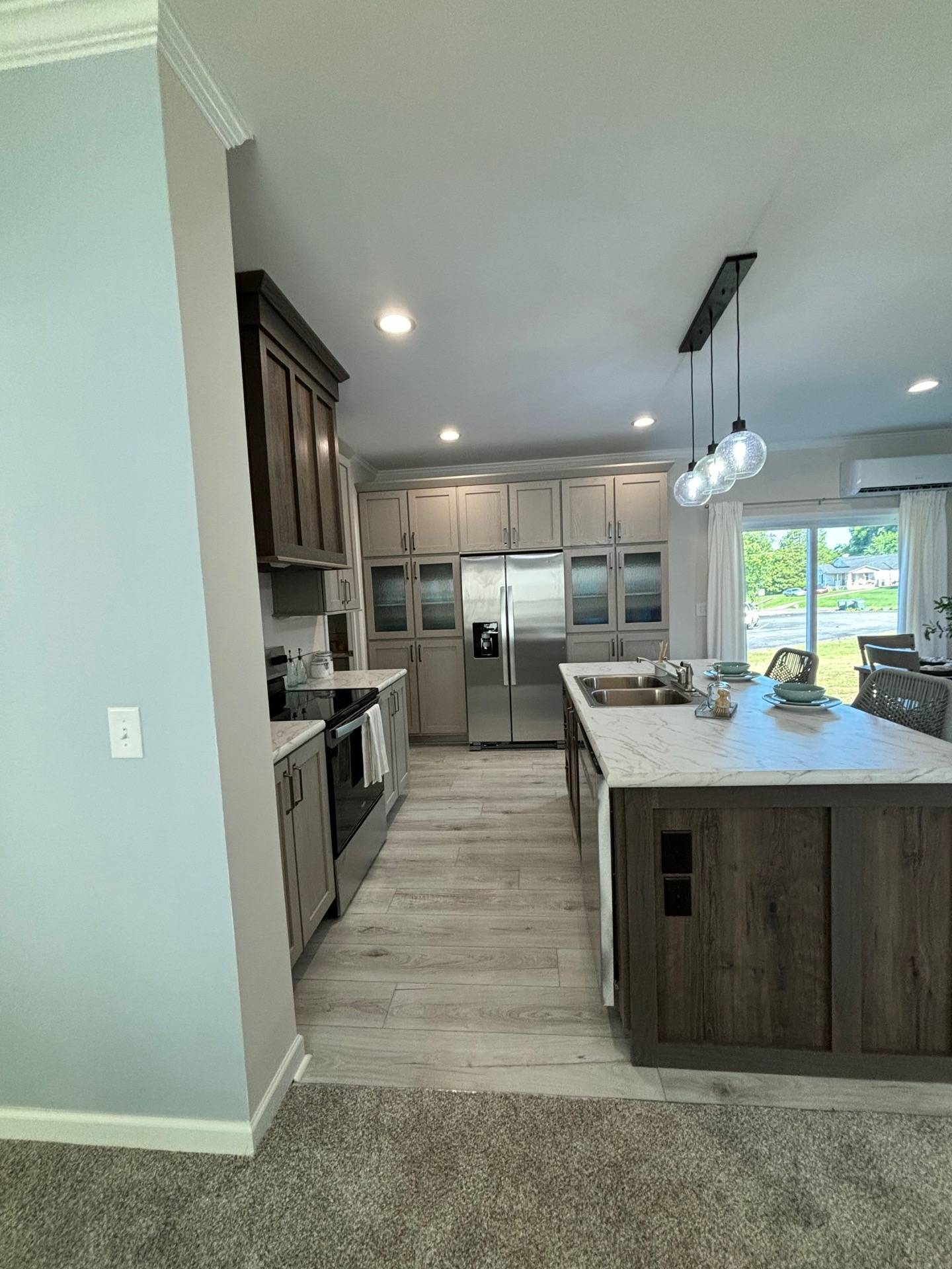 Modern kitchen with dark wood cabinets, stainless steel appliances, and a large marble island. Three pendant lights hang above, creating a bright and welcoming atmosphere. A window offers a view of greenery outside.