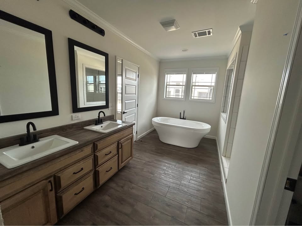 Luxurious spacious master bathroom ensuite in premium modular home featuring double undermount rectangular sinks on rustic reclaimed wood-grain quartz countertop with matte black gooseneck faucets, extra-long white freestanding oval soaking bathtub with black floor-mount faucet, large black-framed rectangular mirrors with integrated black vanity sconce lighting, neutral beige walls with crown molding, dark gray wide-plank luxury vinyl plank flooring, white six-panel doors, tiled shower niche alcove, bright airy contemporary farmhouse-style primary bathroom interior view in new manufactured ranch home