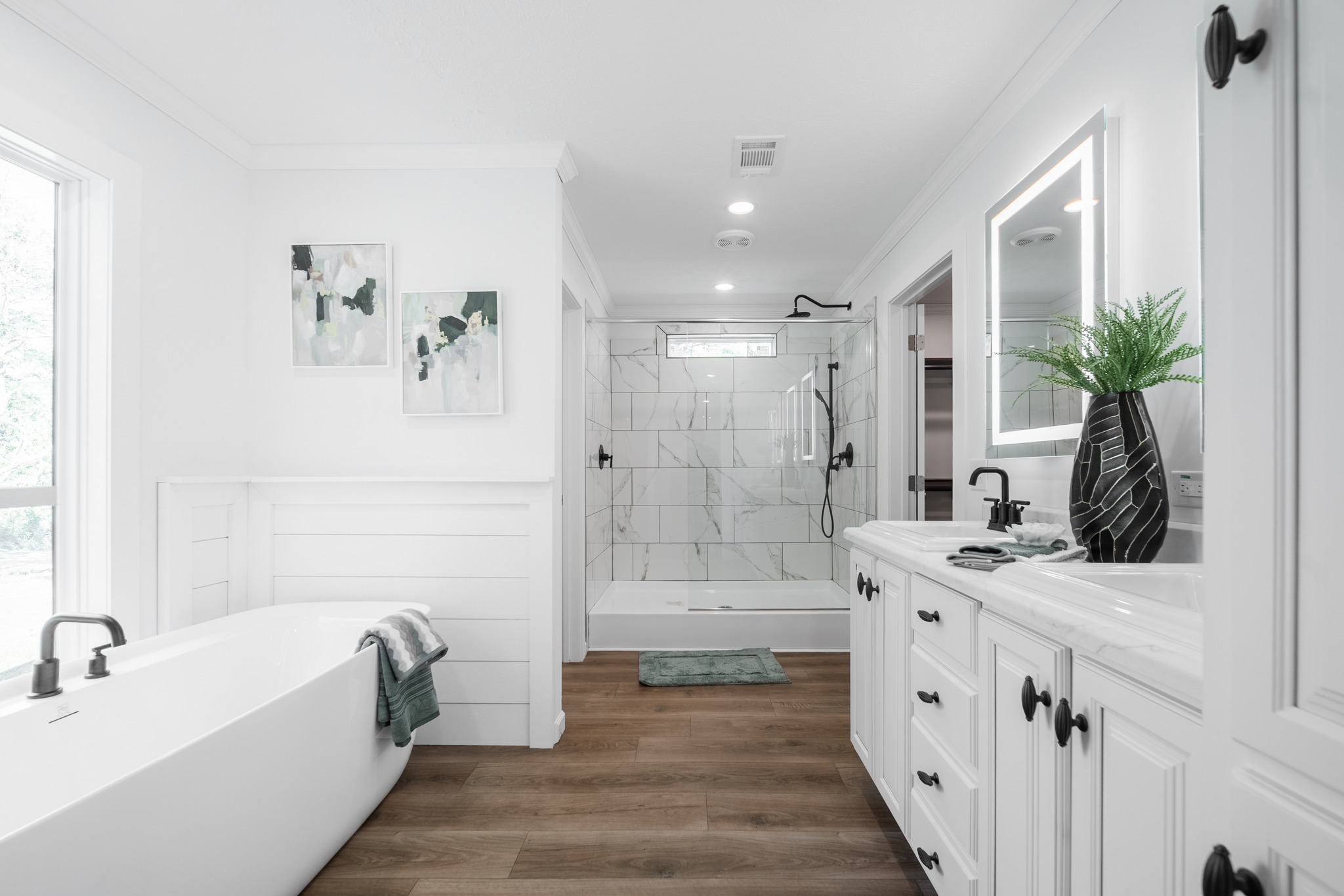 Sleek bathroom with white walls and hardwood floors. Features a freestanding tub, glass shower, double vanity, round mirror, and greenery, creating a serene atmosphere.